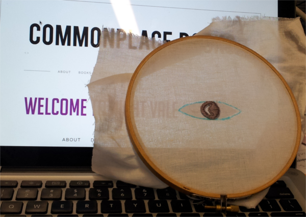 A laptop screen displays the Welcome to Night Vale banner page. An embroidery hoop with an outline of the Night Vale logo is placed in front of the laptop screen, superimposing the embroidery over the logo.